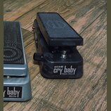 Dvě wah pedály umístěné na dřevěném povrchu. Pravý pedál má černý maskáčový vzor a nápis DIME cry baby FROM HELL. Levý pedál je stříbrný s nápisem cr…