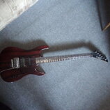 Elektrická kytara s tmavě červeným lakovaným trupem z dřeva s viditnou strukturou zrn. Nástroj má dva humbuckery, jeden tónový potenciometr a přepína…