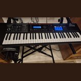 Černý digitální klavír s bílými a černými klávesami, umístěný na černém X-stojanu. Na pravé straně panelu je bílý nápis Roland Juno-DS a sekce PHRASE…