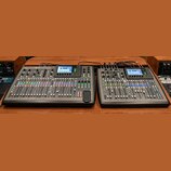 Dvě digitální mixovací konzole černé barvy umístěné vedle sebe na dřevěném povrchu. Na levé konzoli je v horní části viditelné logo a nápis X32. Obě…