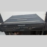 Černý výkonový zesilovač s nápisem AMERICAN AUDIO PROFESSIONAL POWER AMPLIFIER a označením modelu VLP600. Přední panel obsahuje dva ovratné ovládací…
