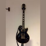 Černá elektrická kytara typu Les Paul se zlatým hardwarem a bílými blokovými vložkami na hmatníku. Na hlavě je viditelné logo Epiphone. Kytara je zav…