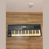Šedý MIDI kontrolér s klávesnicí, faderem a padovými políčkami. Na levé straně je bílý nápis Axiom a v pravém horním rohu logo M-AUDIO. Povrch nástro…