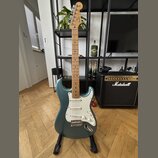 Modrá elektrická kytara typu Stratocaster s bílým pickguardem a světle hnědým hmatníkem. Na hlavě je viditelné logo Fender. Kytara stojí na černém st…