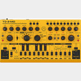 Žlutý analogový basový syntetizátor s nápisem TD-3-MO Modded Out Analog Bass Line Synthesizer. Přístroj obsahuje zahlreiche černé ovládací knofíky, p…