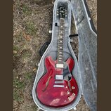 Červená polodublová elektrická kytara v šedém tvrdém pouzdře. Na hlavě hmatníku je bílé logo Gibson. Nástroj má dva humbuckery, černý pickguard a čty…