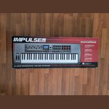 Fotografie znázorňuje krabici s MIDI klaviaturou v barvě stříbrné a černé. Na krabici jsou nápisy IMPULSE 61, novation a text 61 NOTE PROFESSIONAL US…