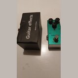 Zelený efekt pedál s nápisy Analog Delay a TRUE BYPASS. Na horní straně jsou tři černé ovládací knoflíky a stisknutý spínač. Vedle pedálu je černá ka…