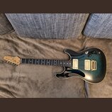 Elektrická kytara s modro-černým přechodovým lakem a krčkem z tmavého dřeva. Na hlavě je viditelné logo Ibanez a nápis RG SERIES. Nástroj má dva humb…