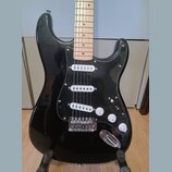 Černá elektrická kytara typu Stratocaster se světlým javorovým hmatníkem. Má černý pickguard a tři bílé snímače typu single-coil. Na ovládacích knofl…