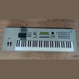 Šedý digitální klavír s bílými a černými klávesami. Na levé straně je nápis YAMAHA a vpravo nápis MOTIF 6. Přístroj obsahuje displej, několik posuvní…