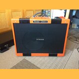 Kytarový kombozesilovač s oranžovým povrchem a černými vertikálními pruhy. Na horní hraně je stříbrný štítek s nápisem Fender - Deluxe a uprostřed če…