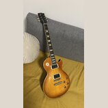 Elektrická kytara s trupem v žlutě-hnědém sunburst provedení a tmavým hnedým hmatníkem s trapezoidními vložkami. Na hlavě je bílý nápis Gibson. Nástr…