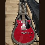 Červená elektrická kytara s černým pickguardem v otevřeném kufru s šedou vnitřní výstuhou. Na hlavě hmatníku je bílý nápis Gretsch. Kytara má dva hum…