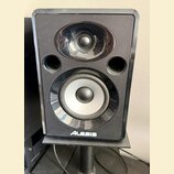 Černý studiový monitor s lesklým povrchem, umístěný na černém stojanu. Na přední straně jsou dva reproduktory a dva kruhové odvětrávací otvory. V dol…