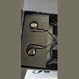 Dva černá šupenová sluchátka s průhledným krytem, pod kterým jsou vidět vnitřní komponenty. Sluchátka jsou umístěna v černém pěnovém vložení uvnitř č…