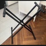 Černý kovový skládací stojan v kształtu X. Povrch je matně černý, s plastovými koncovkami na nohách a horních hranách. Na stojanu nejsou viditelné žá…