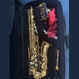 Zlatý saxofon v černém pouzdře. Na korunu nástroje je vyryto "YAMAHA" a pod ním text "Custom Z edition". V pouzdře je umístěna krabička s nápisy "ZZ"…
