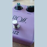 Fialový efekt pedál s nápisem BOSS a symbolem ™. Na horní straně jsou dva černé oválné ovladače s popisky LEVEL a FUZZ. V dolní části je vidět část n…