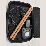 V černém přepravním pouzdře se nacházejí dvě dřevěné paličky s nápisem AeroBand, černý kabel s jack konektory a malý černý oválný modul. Na modulu je…