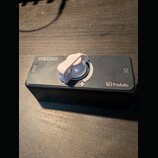 Černý efekt pedál s bílým ovládacím knoflíkem. Na horní ploše jsou nápisy VOLUME, OUT, IN a symbol +. V pravém dolním rohu je logo MH pedals. Povrch…