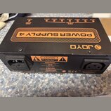 Černý napájecí zdroj s oranžovými prvky a nápisem POWER SUPPLY 4. Na horní straně je viditelné logo JOYO a řada výstupů s označením mA. Na zadní stra…