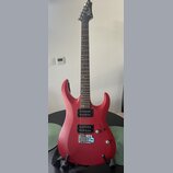 Červená elektrická kytara s matným povrchem a tmavým hnedým hmatníkem. Na hlavě je bílý nápis Cort. Nástroj má dva černá humbuckery, jeden ovládací k…