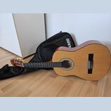 Akustická kytara s přírodním dřevěným povrchem korpusu a hnědým kytarovým hmatníkem. Nástroj leží na dřevěné podlaze vedle černého obalu, na kterém j…