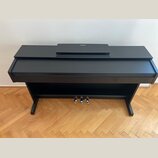 Černý digitální piano v şeklinde nábytkového kusu s uzavřenou klaviaturní víkem. Na horní desce je položen černý krycí prachový kryt s bílým logem YA…