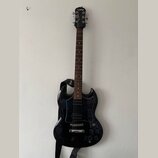Černá elektrická kytara s tmavým hnedým hmatníkem a černým pickguardem. Na hlavě nástroje je bílý nápis Epiphone. Kytara má připevněný černý popruh s…