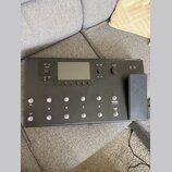 Černý kytarový procesor s displejem a ovládacími prvky. Na panelu jsou nápisy PRESET, VIEW, VOLUME, MODE HOLD TO EDIT a TAP TUNER, dále označení tlač…