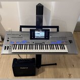 Šedý digitální klavír s logem YAMAHA na horní části a označením Genos na pravé straně. Nástroj je umístěn na černém stojanu a nad ním je průhledlá hu…