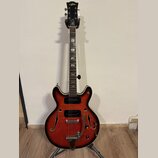 Polodupatá elektrická kytara s červeným sunburst povrchem a bílým bindingem. Na hlavě je zlatý nápis Aria, na hmatníku jsou vidět vložky z perlemi. N…