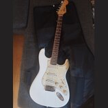 Bílá elektrická kytara s perleťovým pickguardem a hnedým hmatníkem. Na hlavě je nápis Squier STRAT. Nástroj leží na černém softcasu. Povrch kytary vy…