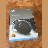 Fotografie znázorňuje krabici od sluchátek. Na šedém povrchu krabice je bílé logo Sennheiser, nápis SENNHEISER a modelové označení HD 600. V levém do…