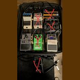 V černé brašně je několik kytarových efektů a příslušenství. Jsou zde pedály značky BOSS s nápisy Digital Delay DD-3, Equalizer GE-7 a Chromatic Tune…