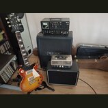 Elektrická kytara s trupem v barevném přechodu od žluté po červenou a bílým pickguardem. Na hlavě kytary je nápis Gibson a La Paul. Vedle ní stojí če…