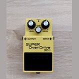 Žlutý efekt pedál s nápisy SUPER OverDrive a SD-1. Na horní části jsou tři stříbrné ovládací knoflíky s popisky LEVEL, TONE a DRIVE a červená kontrol…