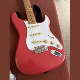 Červená elektrická kytara typu Stratocaster s bílým pickguardem a javorovým hmatníkem. Nástroj má tři bílé snímače, jeden přepínač a tři otočné poten…