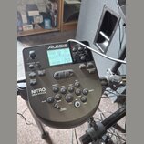 Šedý elektronický bicí modul značky Alesis s nápisem NITRO DRUM MODULE. Zařízení má digitální displej, několik ovládacích tlačítek a volič hlasitosti…
