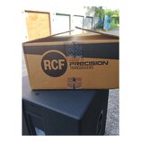 Kartonová krabice s nápisy RCF PRECISION TRANSDUCERS položená na černém reproduktorovém boxu. Krabice je z hnědého kartonu s černým pruhem a je slepe…