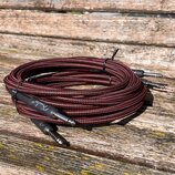 Svinutý instrumentální kabel s červeně a černě pleteným povrchem. Na jednom z černých konektorů je bílý nápis "Fender®". Kabel je stahován černým pla…