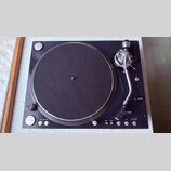 Černý gramofon s kulatým talířem potaženým tmavě šedou plstí a stříbrným vlnitým okrajem. Na raménku je viditelné logo "aton" s modrým symbolem. Ovlá…