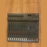 Šedý mixovací pult značky YAMAHA, model Mx12/4. Horní panel obsahuje vstupy, konektory a ovládací prvky s nápisy jako INPUT, GAIN, HIGH, LOW, MON a E…