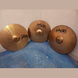 Tři mosazné činely opřené o bílou zeď na modré podlaze. Nejlevý činel vlevo nese nápisy paiste PST II a 18" CRASH RIDE. Prostřední činel má nápisy pA…
