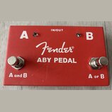 Červený kovový efekt pedál s bílými nápisy. Uprostřed je logo Fender® a text ABY PEDAL, v horní části nápis IN/OUT. Na levé straně jsou označení A a…