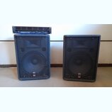 Dva černé reproduktorové boxy s kovovými ochrannými mřížkami. Na spodní části obou boxů je malý štítek s nápisy „SOUND SYSTEM“ a „PROEL“. Na levém re…