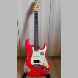 Červená elektrická kytara typu Stratocaster s bílým pickguardem a hnědým hmatníkem. Na hlavě je vidět nápis "Squier". Tělo nástroje vykazuje výrazné…