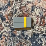 Fotografie zobrazuje malý, obdélníkový elektronický modul nebo kartu v šedé barvě s výrazným žlutým svislým pruhem. V dolní části jsou vidět zlaté ko…