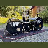 Černá bicí souprava složená z dvou basových bubnů, jednoho tom-tomu a jednoho floor tomu. Na předních blanách basových bubnů je bílý nápis METAL FACT…